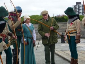 Robert Hardy in a flat cap shows a bow to historical re-enactment people in Mediaeval costume.