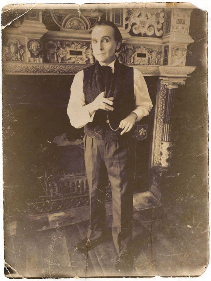 Dressed in mid-19th C costume, Jonathan Goodwin stands in front of an elaborate carved stone fireplace.